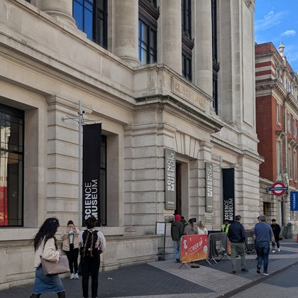 People walking in front of the Science Museum entrance in London, with banners and a red underground sign visible.