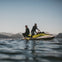 Two men riding a yellow and white jet ski on a calm body of water with mountains visible in the distance under a clear sky.