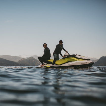 Two men riding a yellow and white jet ski on a calm body of water with mountains visible in the distance under a clear sky.