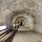 A rocky tunnel with fluorescent lights on the walls and pipes running along the floor, leading to a door at the end.