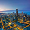 Skyline view of a city at dusk with illuminated skyscrapers and streets, and a large body of water in the background.