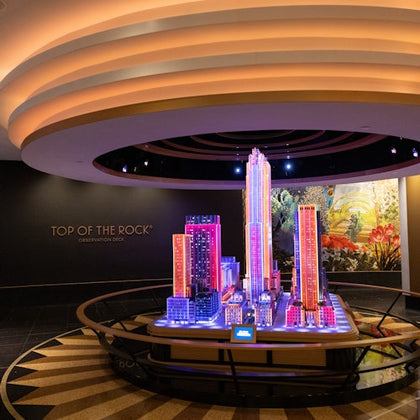 A colorful model of a city skyline displayed indoors under a circular ceiling, labeled "Top of the Rock Observation Deck."