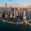 A helicopter flies over the southern tip of Manhattan, with tall skyscrapers and One World Trade Center visible in the background.