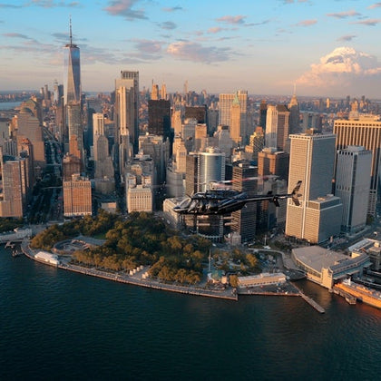 A helicopter flying over lower Manhattan with skyscrapers in the background and the waterfront in the foreground during sunset.