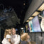 A group of people observe a museum exhibit featuring space exploration displays and a satellite replica in the background.