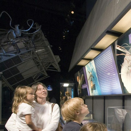 A group of people observe a museum exhibit featuring space exploration displays and a satellite replica in the background.