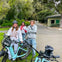 Three people standing outdoors with bicycles, surrounded by greenery and a paved path. A building is visible in the background.