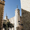 Stone towers and walls of a historic building under a clear blue sky, flanked by trees and a vintage lamp post.