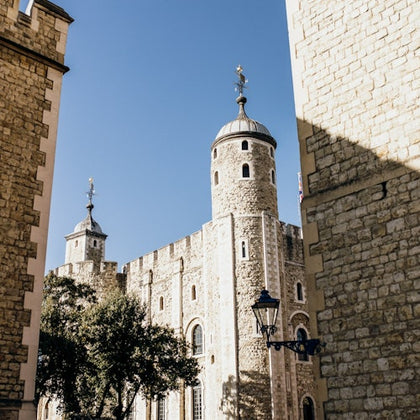 Stone towers and walls of a historic building under a clear blue sky, flanked by trees and a vintage lamp post.
