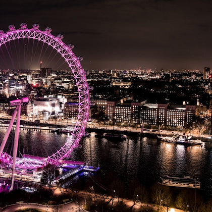 A brightly lit Ferris wheel by a river in a cityscape at night with illuminated buildings and boats on the water.