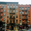 Three adjacent multi-story brick buildings with fire escapes, ground-floor shops, and a street with parked cars.