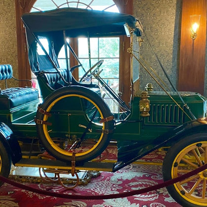 A vintage green automobile with yellow accents displayed in a room with ornate decor and a window in the background.
