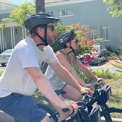 Two people wearing helmets and sunglasses ride bicycles on a sunny street, with a house and flowering bushes in the background.