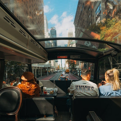 People seated in a bus with a glass roof, viewing a city street with tall buildings, traffic, and a blue sky.