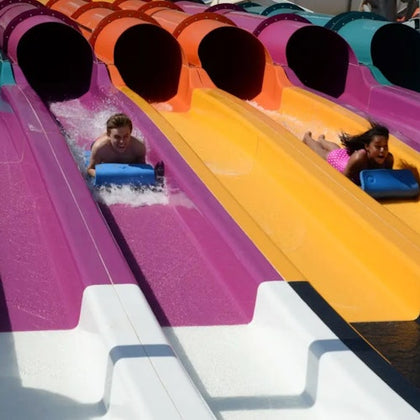 Two people sliding down colorful water slides on blue mats at a water park.