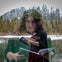 A woman, superimposed with a forest background, reads a book, holding another. Snowy trees and a lake appear behind her.