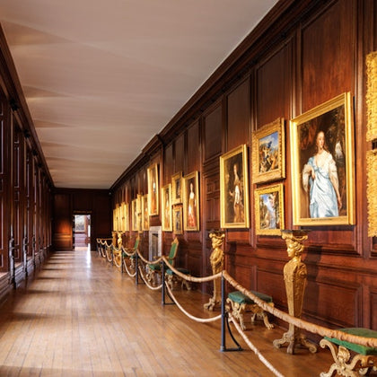 A long gallery with wooden paneling featuring framed paintings, ornate benches, and a roped-off walkway.