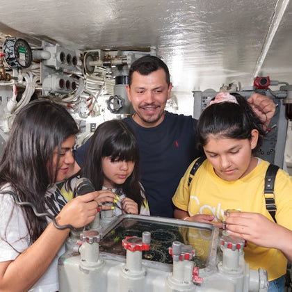 A man and three children inside a submarine, interacting with equipment and controls in a confined technical space.