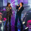 Two performers singing on a lit stage surrounded by an audience and colorful lights.