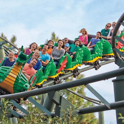 People smiling and raising their hands on a green dragon-themed rollercoaster outdoors under a blue sky.