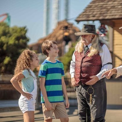 Two children smiling and talking with a man in a cowboy costume outside in a western-themed setting.