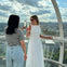 Two women toast with champagne inside a glass observation capsule, with a cityscape and cloudy sky visible in the background.