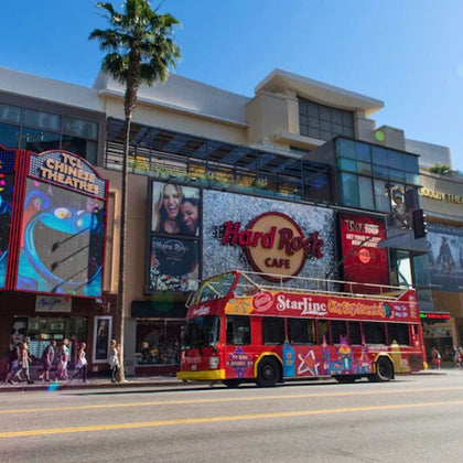 A red double-decker sightseeing bus on a city street, passing by buildings including the Hard Rock Cafe and TCL Chinese Theatre.