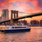 A NY Waterway ferry sails on the river under the Brooklyn Bridge during a vibrant sunset with a city skyline in the background.
