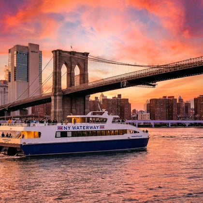 A NY Waterway ferry sails on the river under the Brooklyn Bridge during a vibrant sunset with a city skyline in the background.