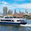 A ferry labeled 'NY Waterway' travels on a river with a city skyline and blue sky in the background.