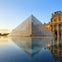 A glass pyramid with a reflection in water in front of a historic building under a clear sky at sunset.