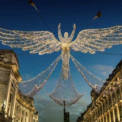 Illuminated angel decoration with outstretched wings hung across a street between two buildings at dusk.