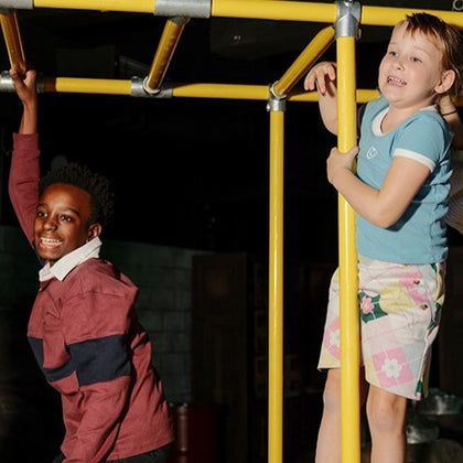 Two children playing on yellow monkey bars indoors, smiling and enjoying the activity.