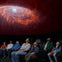 A group of people seated in a planetarium, looking up at a vivid depiction of a galaxy displayed on the dome ceiling.