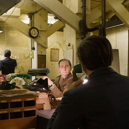 Two men in a dimly lit, cluttered office with industrial decor; one man in military attire, the other in a suit, using a phone.