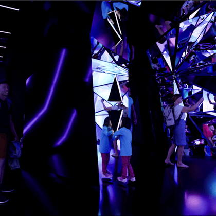 A mirrored room with purple lighting, children and adults interacting with reflective surfaces and enjoying the space.