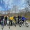 Six people in helmets pose with their bikes on a path near leafless trees and a small body of water under a partly cloudy sky.