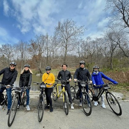 Six people in helmets pose with their bikes on a path near leafless trees and a small body of water under a partly cloudy sky.