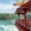 A red cable car suspended over a river with rapids, surrounded by lush green forested cliffs under a partly cloudy sky.