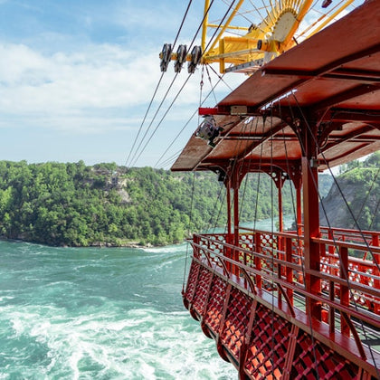 A red cable car suspended over a river with rapids, surrounded by lush green forested cliffs under a partly cloudy sky.