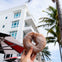A hand holds a powdered donut in front of a white multi-story building with blue windows and palm trees.