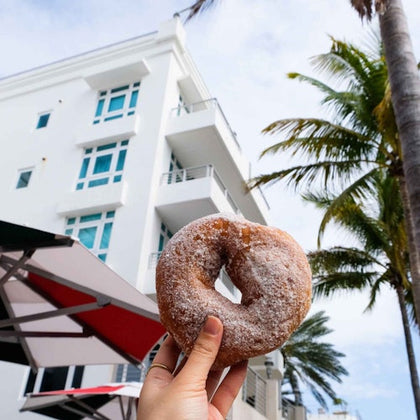 A hand holds a powdered donut in front of a white multi-story building with blue windows and palm trees.