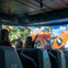 People seated in a simulation theater interacting with a screen showing a vibrant jungle and an orange vehicle.