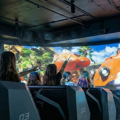 People seated in a simulation theater interacting with a screen showing a vibrant jungle and an orange vehicle.
