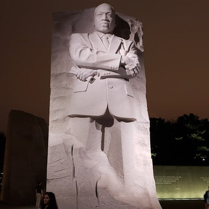 A large stone statue of a man in a suit with crossed arms at night, surrounded by a wall with inscriptions and trees.