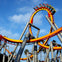 A roller coaster with an orange and yellow loop, carrying people, set against a blue sky with palm trees in the background.