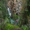 A person walks across a bridge spanning a forested gorge, with a waterfall cascading down the rocky cliffs in the background.