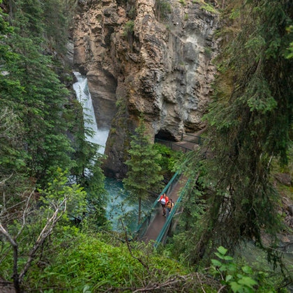 A person walks across a bridge spanning a forested gorge, with a waterfall cascading down the rocky cliffs in the background.