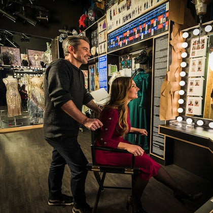 A man and a woman are in a dressing room with illuminated mirrors and costumes displayed in the background.