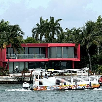 A water taxi passes in front of a modern red house with large windows and palm trees by the waterfront.